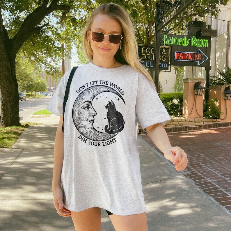 Celestial moon t-shirt featuring a vintage-style crescent moon with a human face and a cosmic black cat sitting on top. Astrology-inspired design with a witchy aesthetic, mystical starry details, and a motivational quote. Soft cotton blend, unisex fit, and available in various sizes. Perfect for astrology lovers, witchcore fashion fans, and those who adore celestial art. Great gift for spiritual souls, moon phase enthusiasts, and black cat lovers.