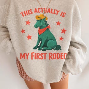 A person wearing a grey sweatshirt with a graphic of a green horse and the text "This actually is my first rodeo" printed on it.