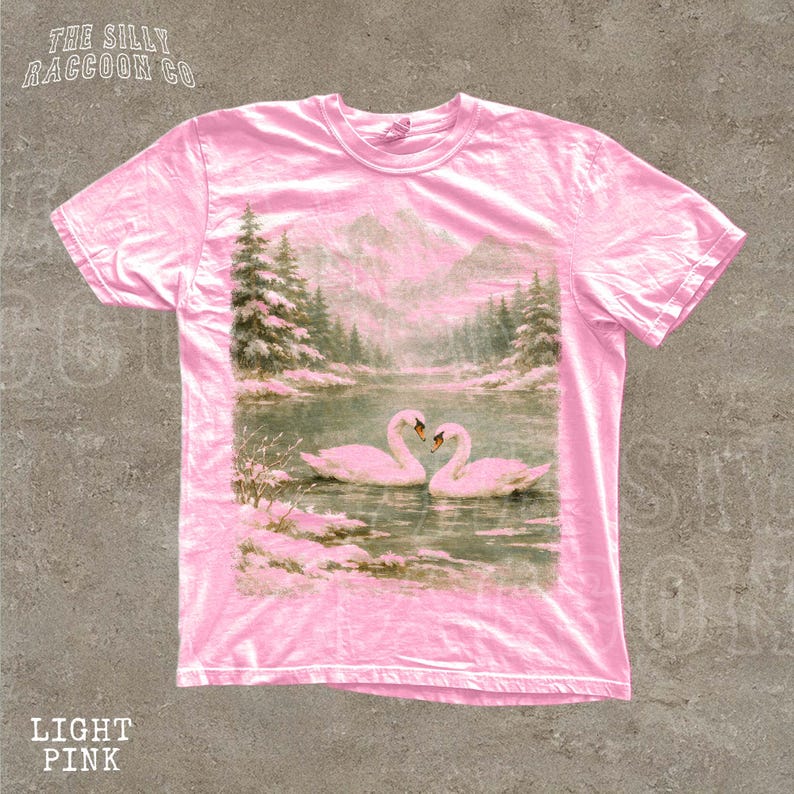 A pink t-shirt with a printed image of two swans swimming in a lake, surrounded by pine trees and mountains.