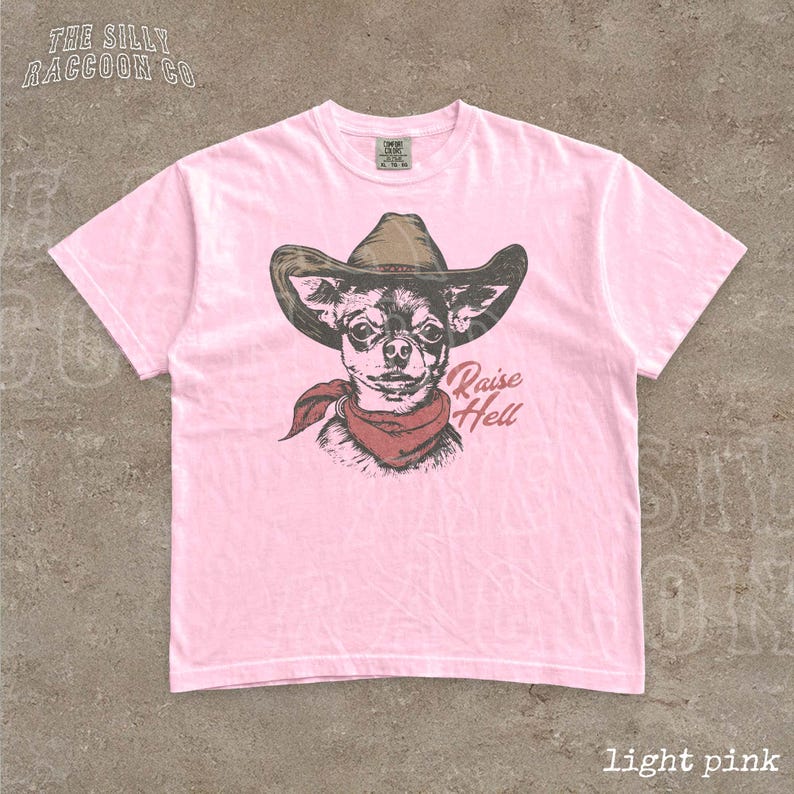 A pink t-shirt with a graphic of a dog wearing a cowboy hat and a red scarf, along with the text "Raise Hell" printed on it.