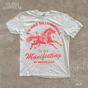 A white t-shirt with a red graphic design depicting a running horse and the text "I'm not delusional, I'm just Manifesting my own reality" printed on it.
