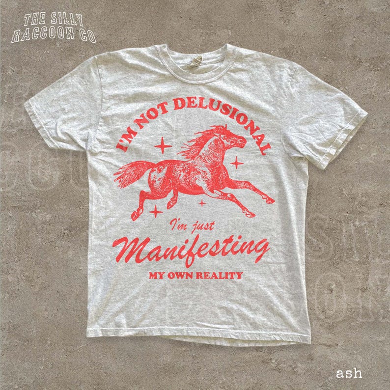 A white t-shirt with a red graphic design depicting a running horse and the text "I'm not delusional, I'm just Manifesting my own reality" printed on it.