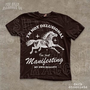 a dark brown t-shirt with a graphic design depicting a horse in mid-gallop, along with the text "I'm not delusional, I'm just Manifesting my own reality" printed on it.