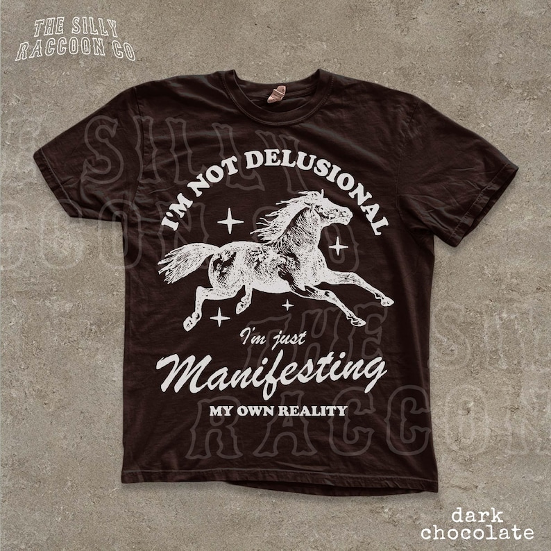 a dark brown t-shirt with a graphic design depicting a horse in mid-gallop, along with the text "I'm not delusional, I'm just Manifesting my own reality" printed on it.