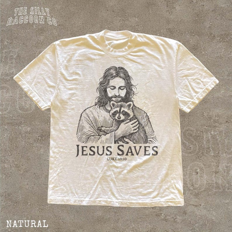A white t-shirt with a graphic design depicting a man holding a raccoon. The text below the graphic says "JESUS SAVES" and "LUKETH 10:10".