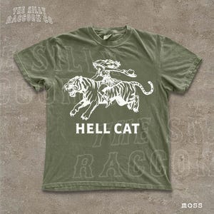 A green t-shirt with a graphic design depicting a tiger and a person riding on its back, along with the text "HELL CAT" printed on it.