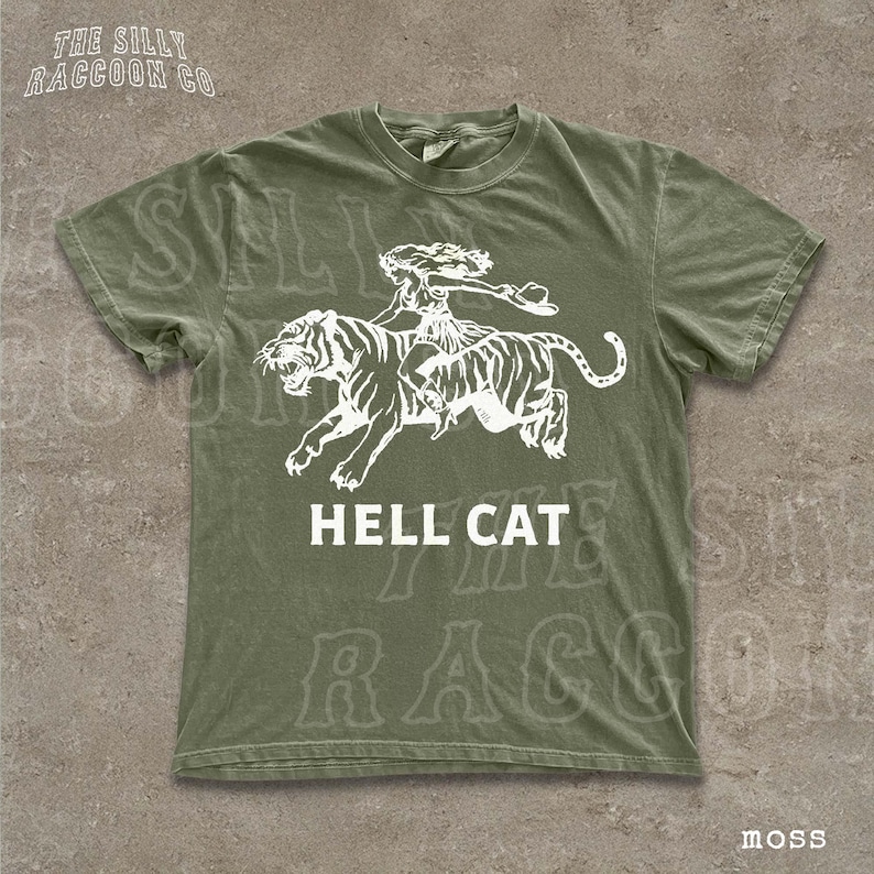 A green t-shirt with a graphic design depicting a tiger and a person riding on its back, along with the text "HELL CAT" printed on it.