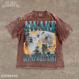 A t-shirt with a graphic design featuring two koalas and the text "AWAKE BUT AT WHAT COST" printed on it.