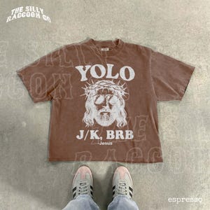 A brown t-shirt with a graphic design and text printed on it, along with a pair of sneakers and a person's feet.