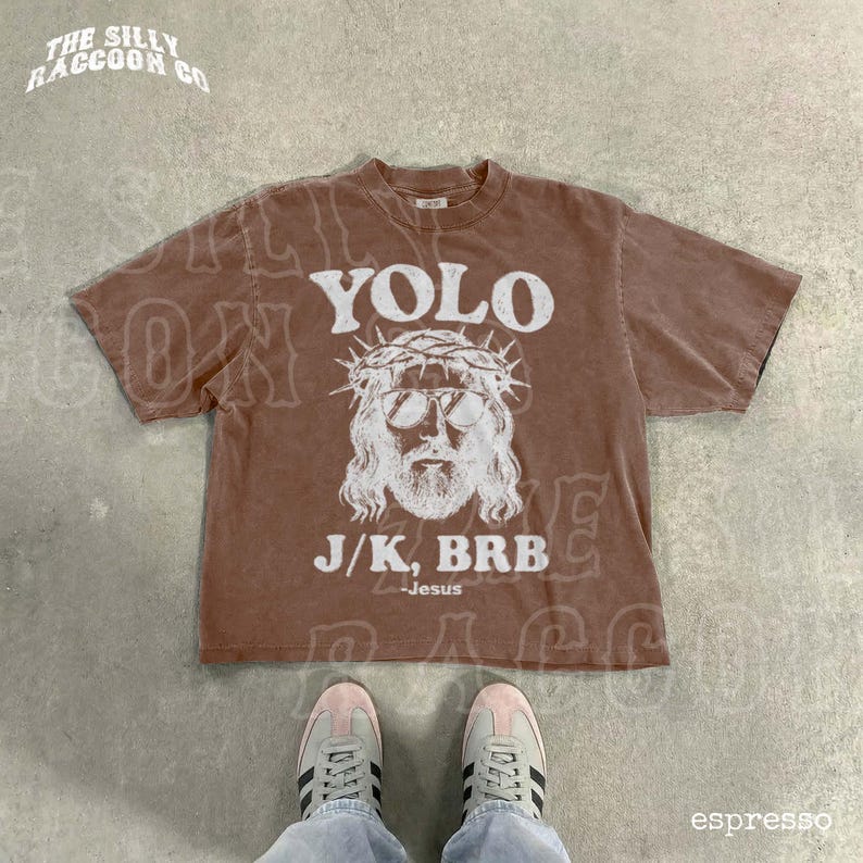 A brown t-shirt with a graphic design and text printed on it, along with a pair of sneakers and a person's feet.