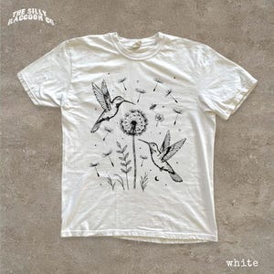 A white t-shirt with a black and white illustration of two hummingbirds hovering over a dandelion surrounded by flowers and plants.