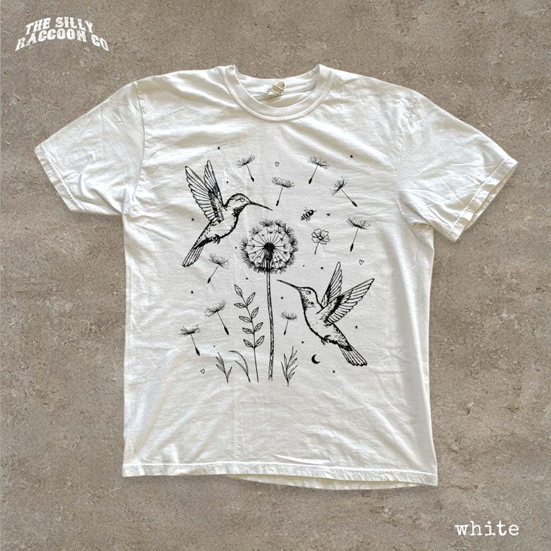 A white t-shirt with a black and white illustration of two hummingbirds hovering over a dandelion surrounded by flowers and plants.