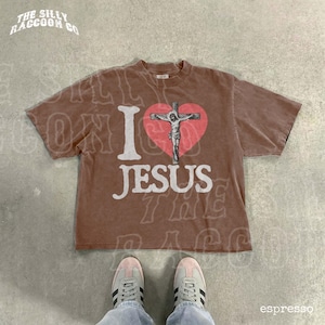 A brown t-shirt with a graphic of a cross and the text "I I J E S" printed on it, along with a heart symbol. The t-shirt is laid out on a concrete floor, and a pair of sneakers is visible underneath it.