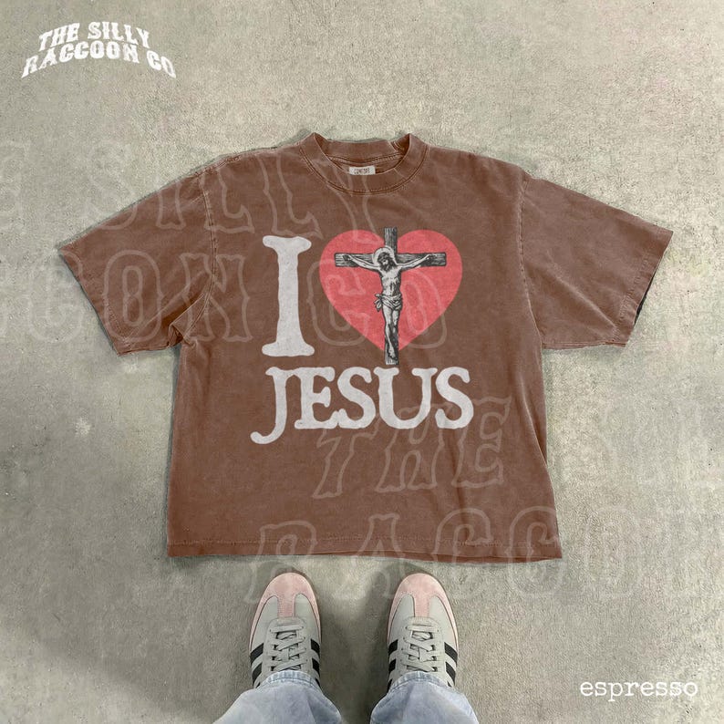 A brown t-shirt with a graphic of a cross and the text "I I J E S" printed on it, along with a heart symbol. The t-shirt is laid out on a concrete floor, and a pair of sneakers is visible underneath it.