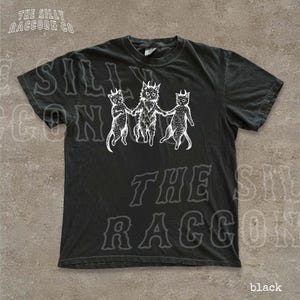 A black t-shirt with a graphic design featuring three cats with horns and a cat-like creature on the left side.