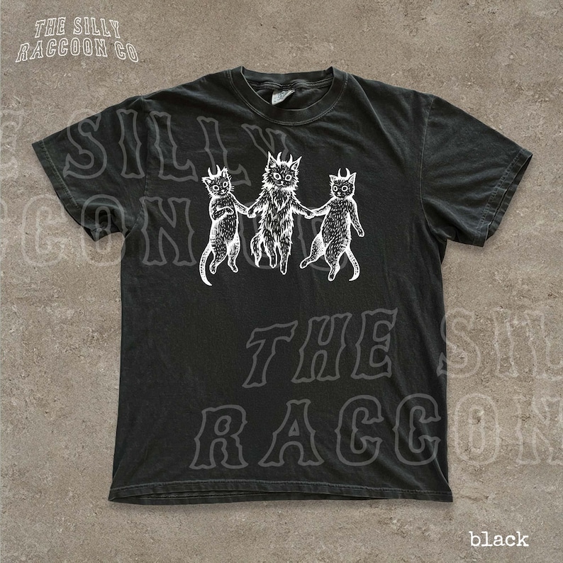 A black t-shirt with a graphic design featuring three cats with horns and a cat-like creature on the left side.