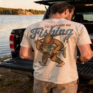 A man standing in front of a truck with a t-shirt featuring a cartoon fish and the text "I'D Rather Be Fishing".
