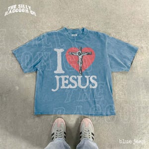 May include: A light blue t-shirt with the text "I Jesus" in white, featuring a red heart with an image of Jesus on the cross. The shirt has a vintage, faded look.