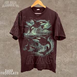 A dark brown t-shirt with a graphic design depicting two orca ( killer whale) leaping out of the water, surrounded by swirling, greenish-blue waves.