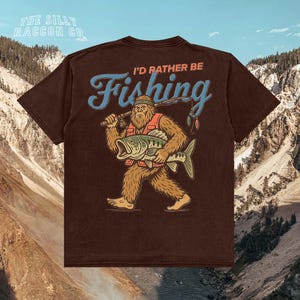 A t-shirt with a graphic design depicting a large, furry creature holding a fish and the text "I'D Rather Be Fishing" against a mountainous background.