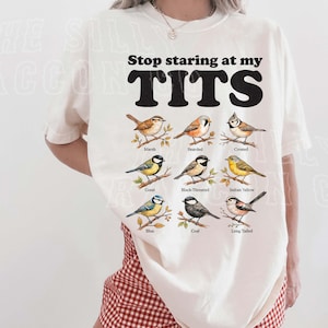 A woman wearing a white t-shirt with a graphic design of various bird species and the text "Stop staring at my TITS" printed on it.
