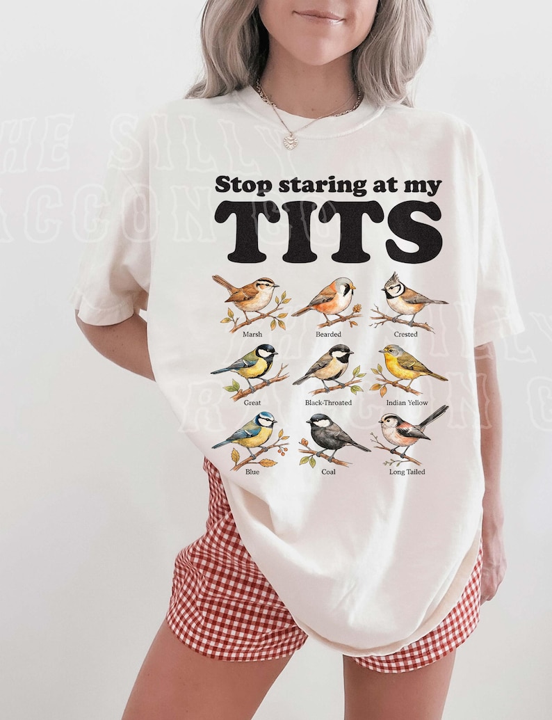 A woman wearing a white t-shirt with a graphic design of various bird species and the text "Stop staring at my TITS" printed on it.