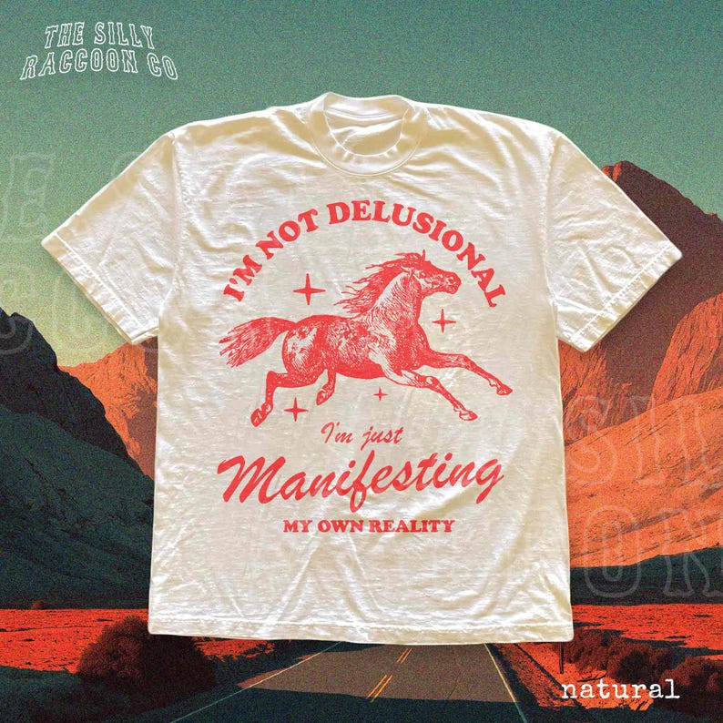 a white t-shirt with a graphic design of a red horse and the text "I'm not delusional, I'm just Manifesting my own reality" printed on it.
