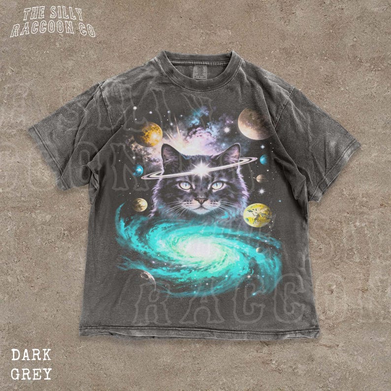 A t-shirt with a graphic design depicting a cat surrounded by a cosmic scene, including planets, stars, and a swirling galaxy.