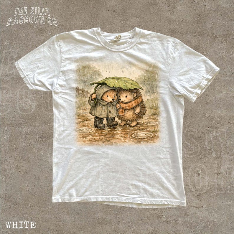 A white t-shirt with a printed illustration of two hedgehog characters standing together under an umbrella.