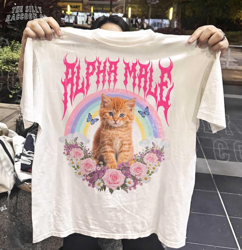 A person is holding up a white t-shirt with a graphic design featuring an orange cat surrounded by flowers and a rainbow.