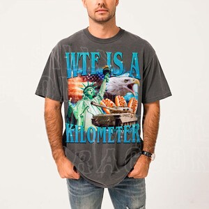 A man wearing a gray t-shirt with a graphic design featuring the Statue of Liberty, an eagle, and a sign that says "WTF IS A KILOMETER".