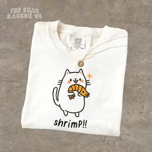 May include: Cream-coloured t-shirt featuring a cartoon cat with a shrimp and the word "shrimp!!" below. The top left corner has the text "THE SILLY RACCOON CO". A gold necklace with a pendant is on the shirt.