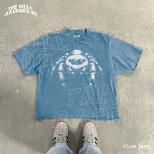 A blue t-shirt with a spider design printed on it, laid out on a concrete floor. A pair of white sneakers is visible underneath the t-shirt.
