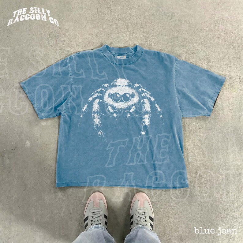 A blue t-shirt with a spider design printed on it, laid out on a concrete floor. A pair of white sneakers is visible underneath the t-shirt.