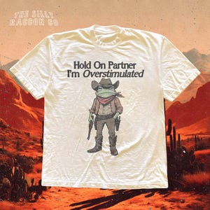 Könnte beinhalten: Weißes T-Shirt mit dem Text "Hold On Partner I'm Overstimulated". Die Grafik zeigt einen Cartoon-Frosch als Cowboy, komplett mit Hut, Halstuch und Pistolen. Das Shirt hat einen Rundhalsausschnitt und kurze Ärmel.
