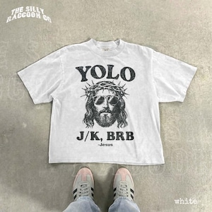 A white t-shirt with a graphic design featuring a man's face wearing sunglasses and a crown, along with the text "YOLO J.K. BRB Jesus". Below the t-shirt, there are a pair of white sneakers.