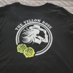 May include: Black t-shirt featuring a white circular design with the text "THE YELLOW ROSE" and a silhouette of a woman wearing a hat. Two yellow roses are below the circle. The shirt is made of a soft material.