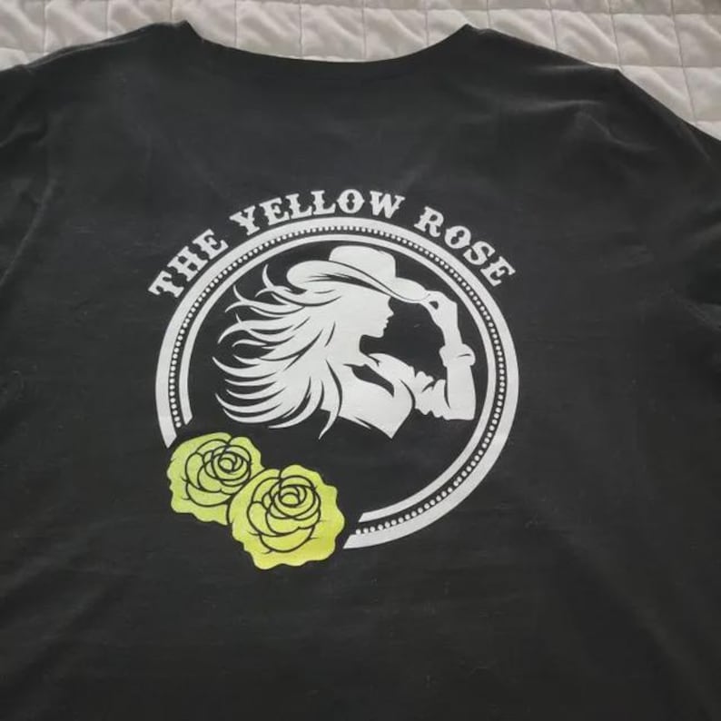 May include: Black t-shirt featuring a white circular design with the text "THE YELLOW ROSE" and a silhouette of a woman wearing a hat. Two yellow roses are below the circle. The shirt is made of a soft material.