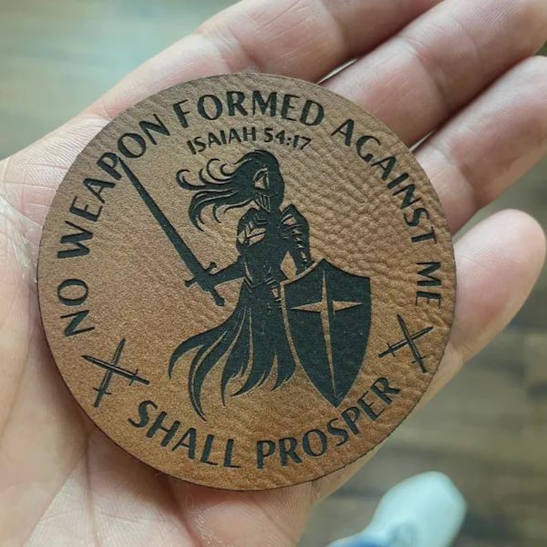 May include: A circular, brown leather coaster featuring the engraved words "NO WEAPON FORMED AGAINST ME SHALL PROSPER" and "ISAIAH 54:17". A black silhouette of a warrior with a sword and shield is in the centre. Two crossed swords are on each side.