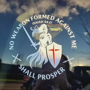 May include: White decal depicting a knight with a sword and shield bearing a red cross. The text states "NO WEAPON FORMED AGAINST ME SHALL PROSPER" and "ISAIAH 54:17". The background is a blurred outdoor scene.