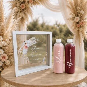 May include: A white square shadow box frame with a floral wreath design and the text Anna & Giacomo 10.10.2027, accompanied by two clear plastic bottles filled with pink and dark red sand, each labeled with the names Anna and Giacomo.