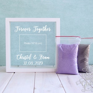 May include: A white framed wedding keepsake with the words "Forever Together" and a photo space. The names "Christel & Bram" and the date "31.08.2019" are also displayed. Two clear bags of colored sand are next to the frame.