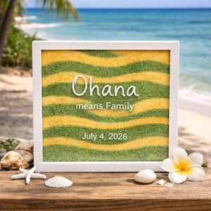 May include: A rectangular white frame containing layered yellow and green sand in a wavy pattern. The text "Ohana means Family" and "July 4, 2026" is printed in white on the glass front. The frame is decorated with a white starfish and a white flower.