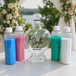 May include: A clear glass heart-shaped decanter with a stopper, featuring the black text The Smiths and the date 08.08.2027. It is surrounded by four clear plastic bottles filled with blue, pink, teal, and white sand for a unity ceremony.