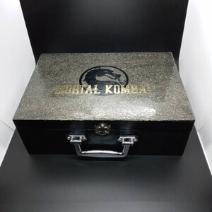Puede incluir: Una caja de madera negra con una manija plateada y un logotipo dorado de Mortal Kombat en la parte superior. La caja tiene una superficie texturizada.