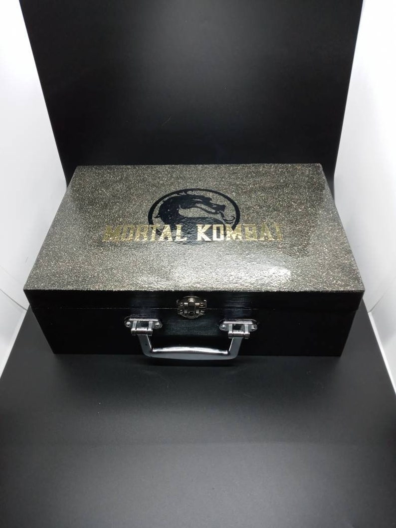 Puede incluir: Una caja de madera negra con una manija plateada y un logotipo dorado de Mortal Kombat en la parte superior. La caja tiene una superficie texturizada.