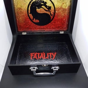 Puede incluir: Caja de madera negra con asa plateada. La tapa est&aacute; abierta y presenta un fondo de purpurina dorada y roja con un logotipo de drag&oacute;n negro. El interior de la caja es negro con la palabra "FATALITY" escrita en rojo.