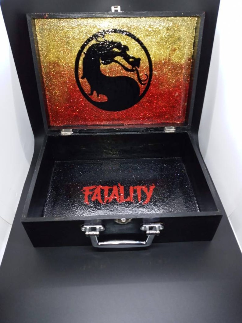 Puede incluir: Caja de madera negra con asa plateada. La tapa est&aacute; abierta y presenta un fondo de purpurina dorada y roja con un logotipo de drag&oacute;n negro. El interior de la caja es negro con la palabra "FATALITY" escrita en rojo.
