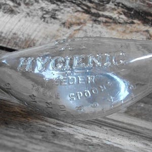 May include: A clear glass baby feeder spoon with the embossed text "HYGIENIC FEEDER SPOONS". The spoon has measurement markings along the side. The spoon is resting on a wooden surface.