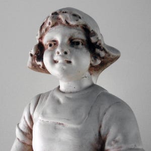 May include: A vintage, off-white ceramic figurine of a young child. The child is wearing a simple outfit and a hat. The figurine has a slightly aged appearance, with subtle brown accents on the hair and hat, adding to its antique charm.